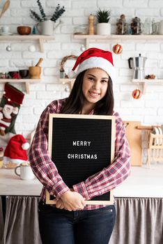 Merry Christmas and Happy New Year. Portrait of a happy beautiful latin woman in santa hat with black letter board with the words Merry Christmas on home kitchen background