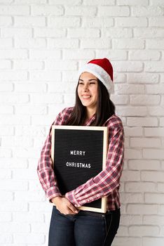 Merry Christmas and Happy New Year. Portrait of a happy beautiful latin woman with letter board with the words Merry Christmas on white brick wall background