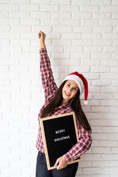 Merry Christmas and Happy New Year. Portrait of a happy beautiful latin woman with letter board with the words Merry Christmas on white brick wall background