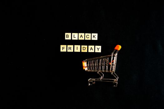 Black Friday written in wooden letters on a black background with a shopping basket, sales, holiday sales, top view, flat layout, price reduction.