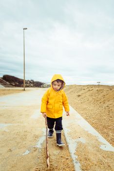 Little boy playing with wood stick, toddler wearing yellow raincoat and gumboots, coastal area