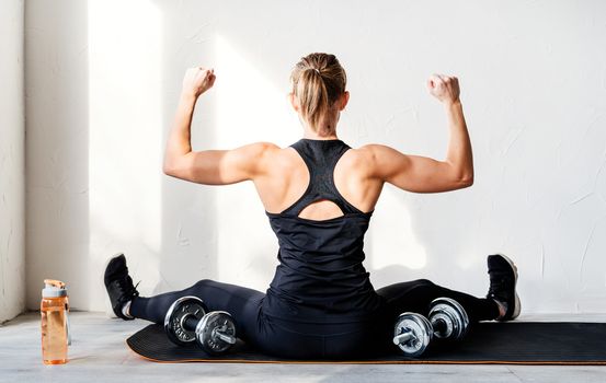 Healthy lifestyle. Sport and fitness. Rear view of young blond woman working out with dumbbells showing her back and arms muscles