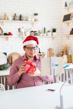 Christmas online greetings. Happy young man in santa hat greeting his friends in video chat or call on tablet sitting in his kitchen