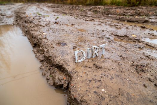 the word dirt composed of silver metal letters on wet clay surface with selective focus, background blur and linear perspective in cloudy autumn day light