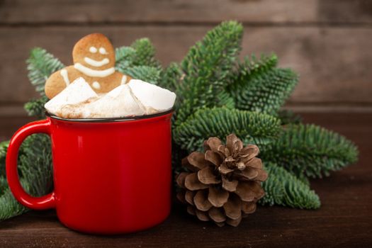 Cocoa hot chocolate in red mug with marshmallows and gingerbread cookie man , fir tree branches and pine cones on dark wooden background