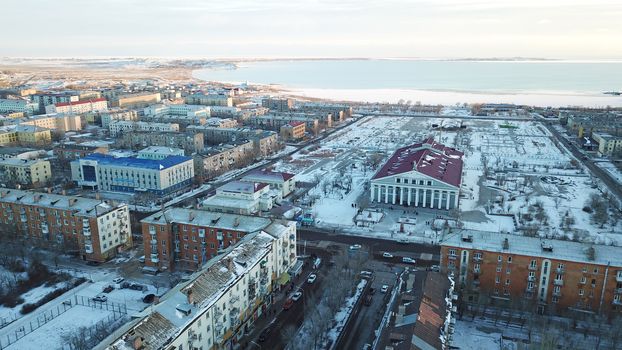 A small town on the shore of lake Balkhash. Top view of houses, cultural buildings, snowy streets, gray roads with cars and a green lake. The lake is partially covered with ice. Soviet city.