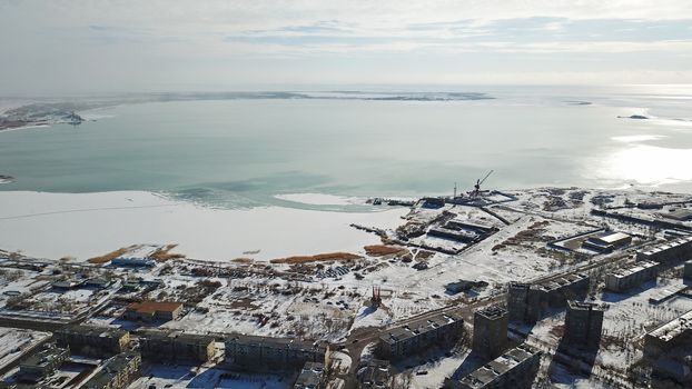 A small town on the shore of lake Balkhash. Top view of houses, cultural buildings, snowy streets, gray roads with cars and a green lake. The lake is partially covered with ice. Soviet city.