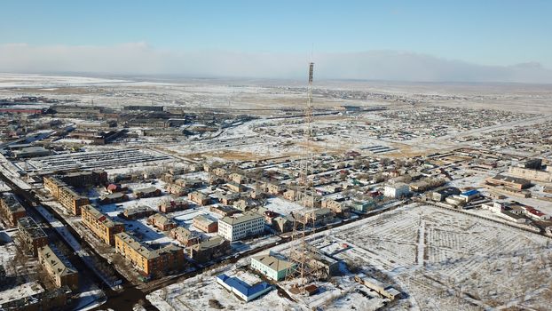 A small town on the shore of lake Balkhash. Top view of houses, cultural buildings, snowy streets, gray roads with cars and a green lake. The lake is partially covered with ice. Soviet city.