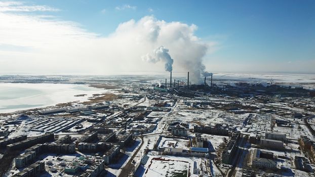 White smoke from the pipes of a factory near the city of Balkhash. Strong emissions in the air. Ecological disaster. Near a small town, houses and a lake. There is snow, blue clear sky and grayness.