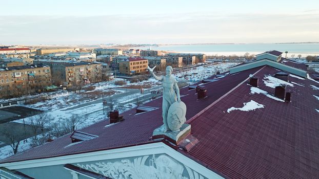 Statue of a woman with a shield on the roof of the Palace. Red tiles on the roof. View of the lake, houses and sunset. The small town of Balkhash in winter.