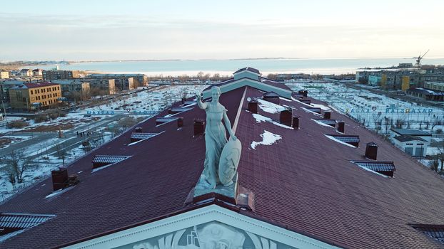 Statue of a woman with a shield on the roof of the Palace. Red tiles on the roof. View of the lake, houses and sunset. The small town of Balkhash in winter.