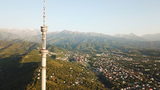 Kok Tobe big TV tower on the green hills of Almaty. View of the mountains, sky, green hills, houses and road. People are walking around the square and alleys below. Houses are visible in the distance