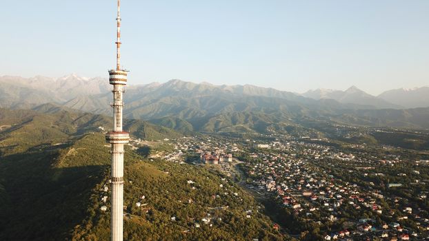 Kok Tobe big TV tower on the green hills of Almaty. View of the mountains, sky, green hills, houses and road. People are walking around the square and alleys below. Houses are visible in the distance