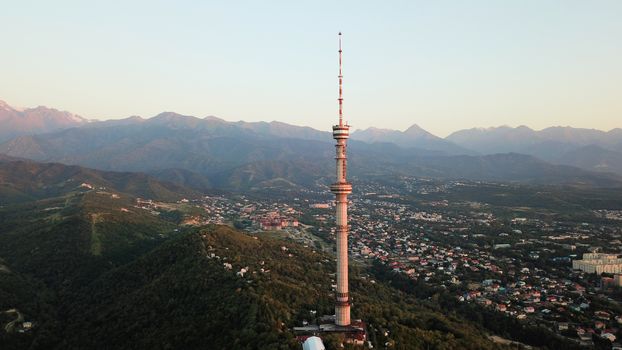 Kok Tobe big TV tower on the green hills of Almaty. View of the mountains, sky, green hills, houses and road. People are walking around the square and alleys below. Houses are visible in the distance