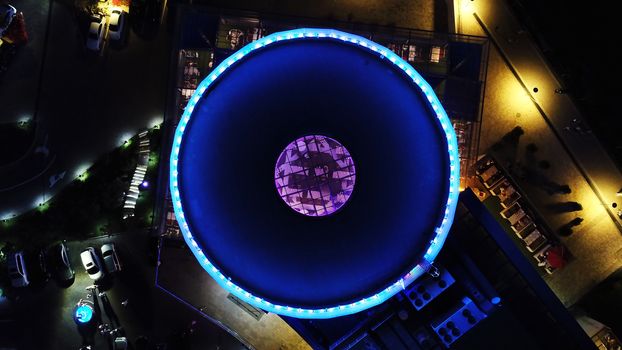 The round roof glows blue in the dark. The view from the top. The roof of the building has transparent ceiling and neon lighting at the edges and in the center. Around the lights and the road. Almaty