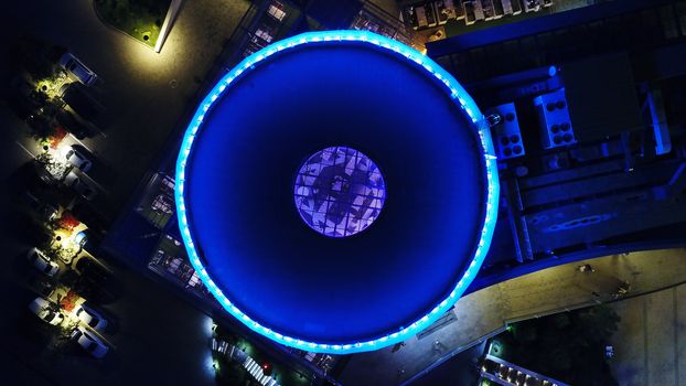 The round roof glows blue in the dark. The view from the top. The roof of the building has transparent ceiling and neon lighting at the edges and in the center. Around the lights and the road. Almaty