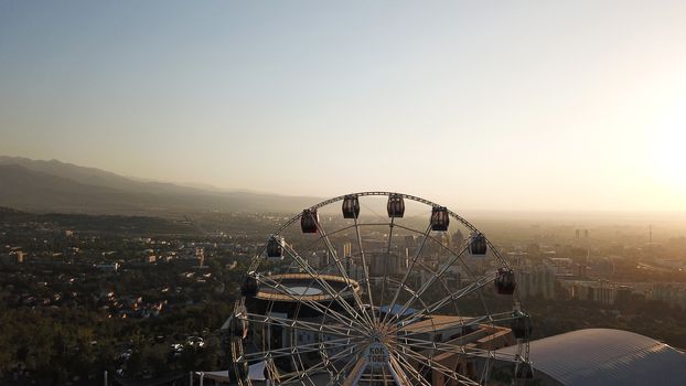 Ferris wheel on the green hill of Kok Tobe at sunset. It offers a view of the city of Almaty, the road, houses and the sky. In places, you can see the city's smog and air pollution. Romantic setting.