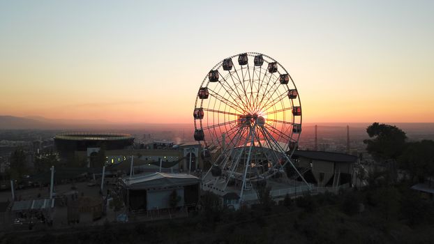 Ferris wheel on the green hill of Kok Tobe at sunset. It offers a view of the city of Almaty, the road, houses and the sky. In places, you can see the city's smog and air pollution. Romantic setting.