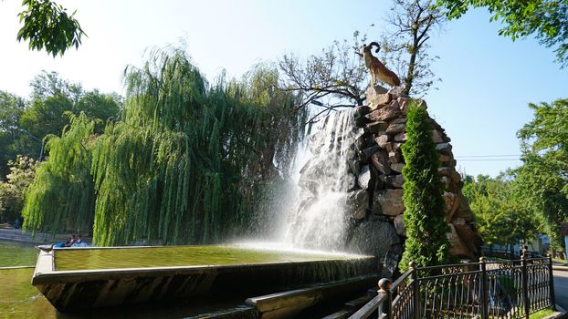 Artificial waterfall with a statue in the city Park of Almaty. Mountain sheep stands on the high stones of the waterfall, water flows into the bowl. Green trees and sky all around.