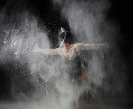 beautiful caucasian woman in a black bodysuit with a sports figure is dancing in a white cloud of flour on a black background