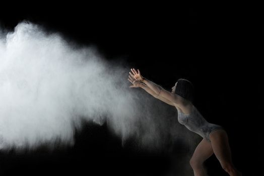 beautiful caucasian woman in a black bodysuit with a sports figure is dancing in a white cloud of flour on a black background, white jet of smoke flies up