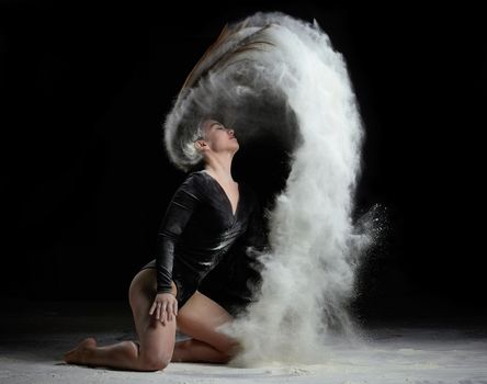 young beautiful caucasian woman with long hair is dressed in a sports black bodysuit and sits on the floor and throwing white flour up, black background