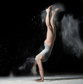young woman with an athletic figure dressed in a black bodysuit stands upside down on her hands, black background with white flying flour, acrobatic exercises