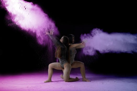 two beautiful young caucasian women in black bodysuits with a sports figure are dancing in a purple-blue cloud of flour on a black background, explosion and expresse in motion