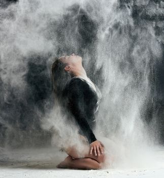 young beautiful caucasian woman with long hair is dressed in a sports black bodysuit and sits on the floor and throwing white flour up, black background