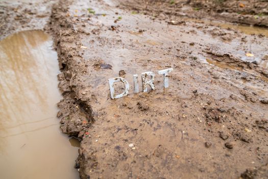 the word dirt composed of silver metal letters on wet clay surface with selective focus, background blur and linear perspective in cloudy autumn day light