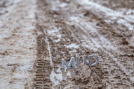 the word mud composed of silver metal letters on wet dirt surface with selective focus, background blur and linear perspective in cloudy autumn day light