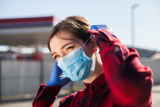 Young woman adjusting protective face mask,standing on petrol station parking lot, Coronavirus COVID-19 corona virus disease global pandemic outbreak crisis, people wearing medical equipment in public