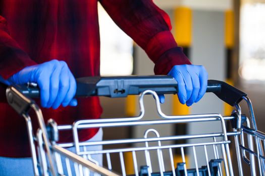 Person wearing blue protective surgical latex rubber gloves pushing shopping cart trolley, stocking supplies, panic buying and hoarding due to global Coronavirus COVID-19 pandemic crisis outbreak