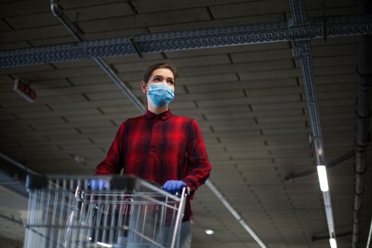 Caucasian woman pushing shopping cart in supermarket underground garage wearing gloves and mask, worried and anxious, panic buying and stocking supplies due to Coronavirus COVID-19 pandemic crisis