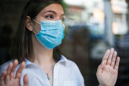 Beautiful young woman wearing face mask,looking through window outside,hands against glass,Coronavirus COVID-19 global pandemic crisis,stay at home social distance order,prevention and protection 
