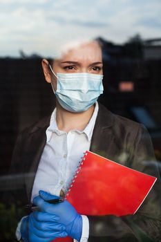 Young anxious caucasian business woman looking through office window,wearing smart clothes,protective gloves,face mask,preventive protection measures at work in UK due to Coronavirus COVID-19 pandemic
