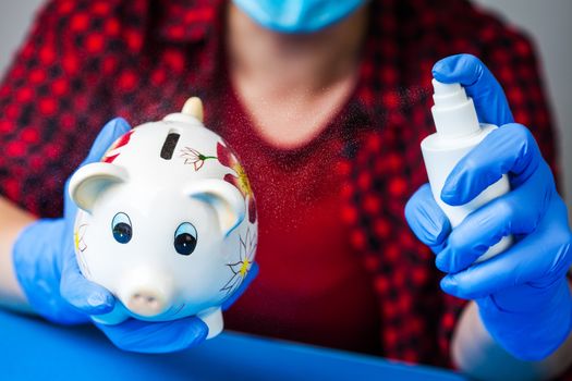 Coronavirus COVID-19 corona virus disease global pandemic outbreak,person holding piggy bank wearing blue surgical protective gloves,disinfecting it with antibacterial sanitizer spray,economy crisis 