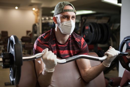 Young caucasian man working out in an indoor gym, barbell bicep curl exercise, wearing protective face mask, sport & fitness during COVID-19 pandemic, health and safety measures and social distancing