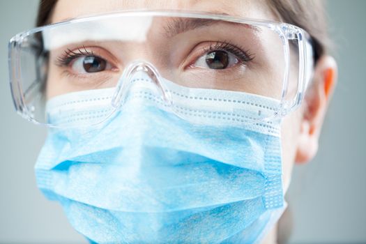Closeup of a worried female NHS UK doctor or lab scientist wearing PPE safety glasses & blue face mask,COVID-19 corona virus disease global pandemic outbreak,uncertainty anxiety & lost hope concept