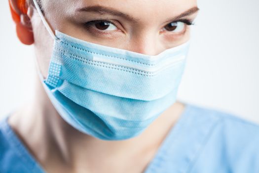 Young woman closeup detail headshot,wearing blue scrubs & protective face mask,attractive beautiful caucasian female doctor staring at camera,pretty eyes giving significant look,Coronavirus COVID-19