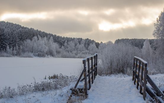 Wooden entrance to the forest near the frozen lake near the forest, all covered in snow. Dramatic sky with clouds over the winter forest and lake. Winter and frosty nature.