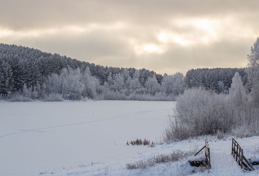 Wooden entrance to the forest near the frozen lake near the forest, all covered in snow. Dramatic sky with clouds over the winter forest and lake. Winter and frosty nature.