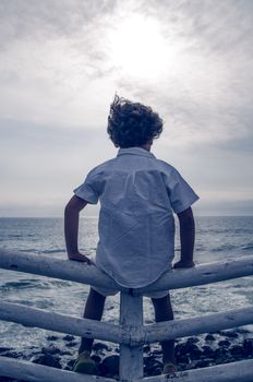 Boy with white clothes sitting on a fence facing the sea