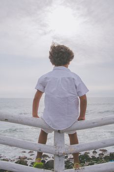 Boy with white clothes sitting on a fence facing the sea