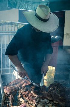 Young white chef with black apron and hat standing near the brazier with coal. Man cooks pork meat outdoor.