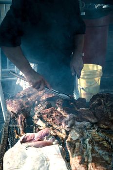 Young white chef with black apron and hat standing near the brazier with coal. Man cooks pork meat outdoor.