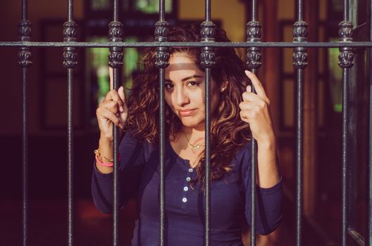 Young girl closed behind bars, network, as in prison. looking sexy with serious and smiling look