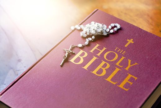 A rosary of white beads with a silver crucifix above the cover of the closed holy bible book. Religion and hope. Religious objects for praying