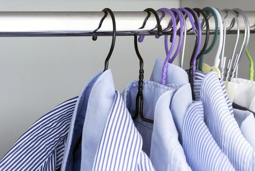 Group of light blue striped shirts hanging on the rod of a white wardrobe. Men's fashion and classic clothing.