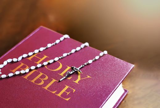 A rosary of white beads with a silver crucifix above the cover of the closed holy bible book. Religion and hope. Religious objects for praying